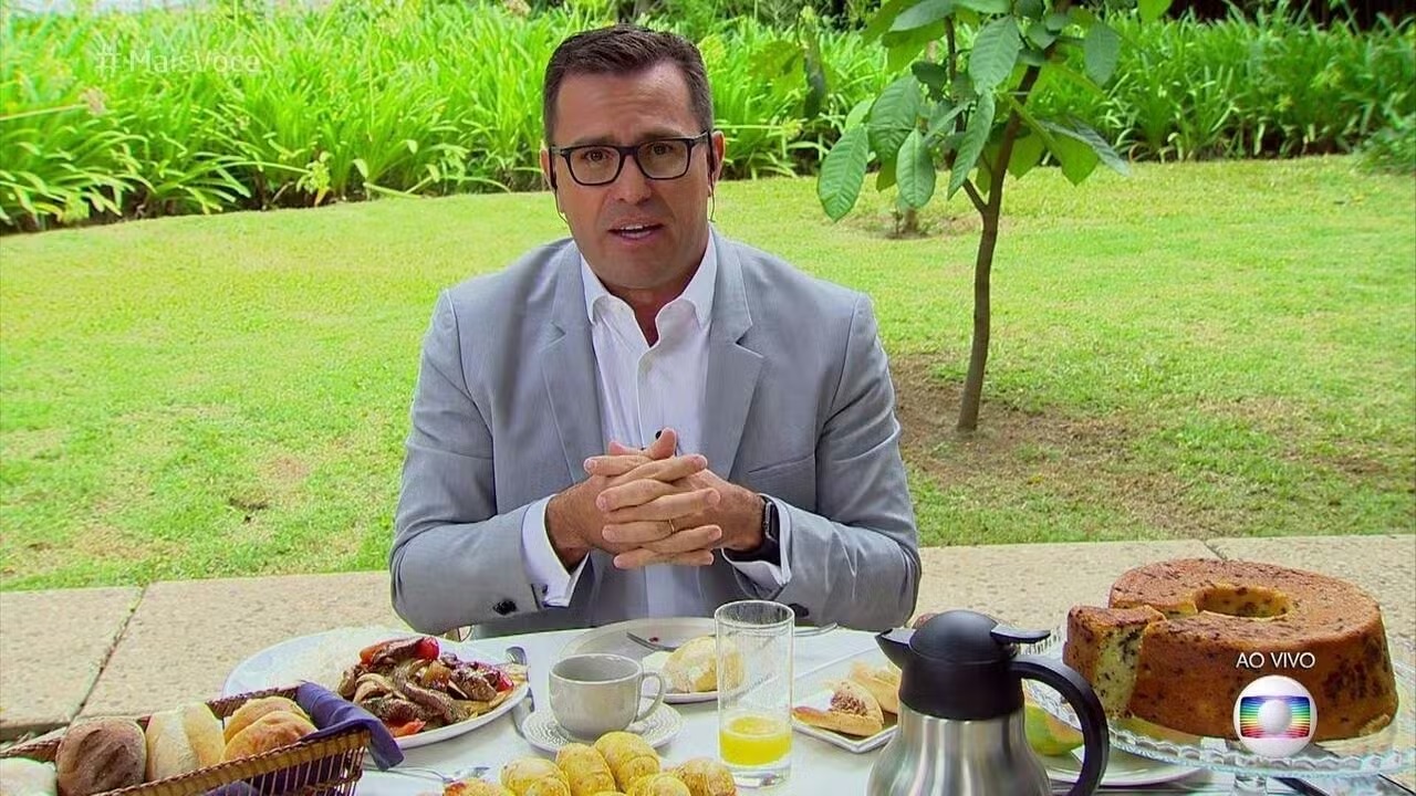 Rodrigo Bocardi sentado perto de mesa de café da manhã, sério, de óculos, falando