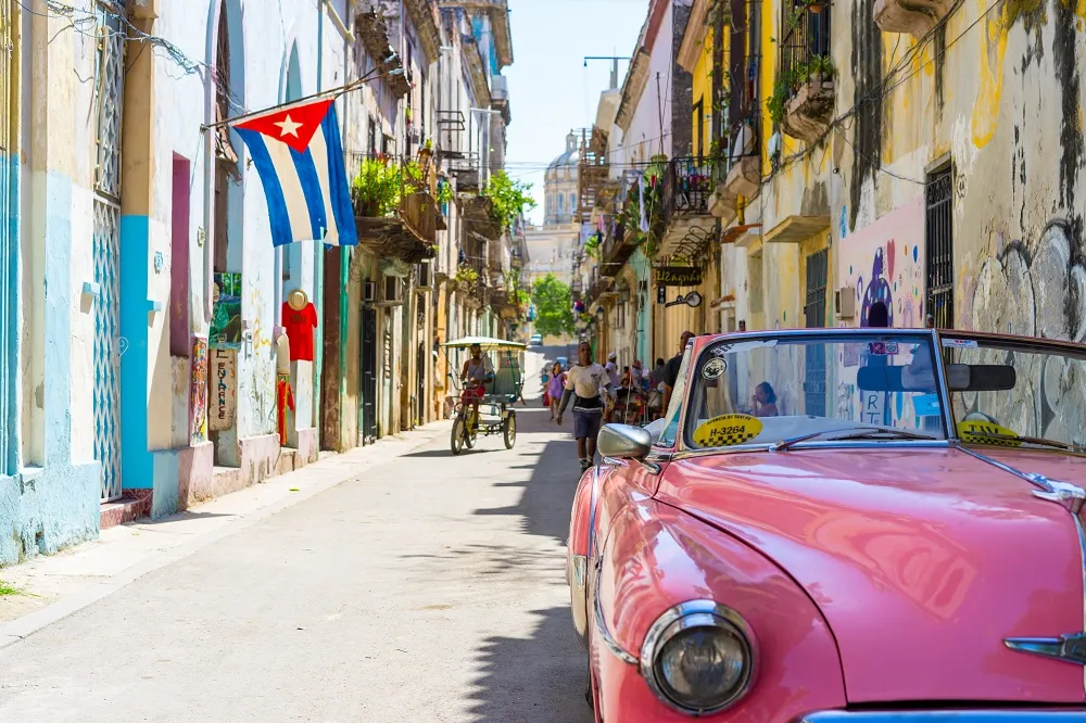 Paisagem cubana com carro antigo cor de rosa e pessoas caminhando na rua, com bandeira cubana aparecendo