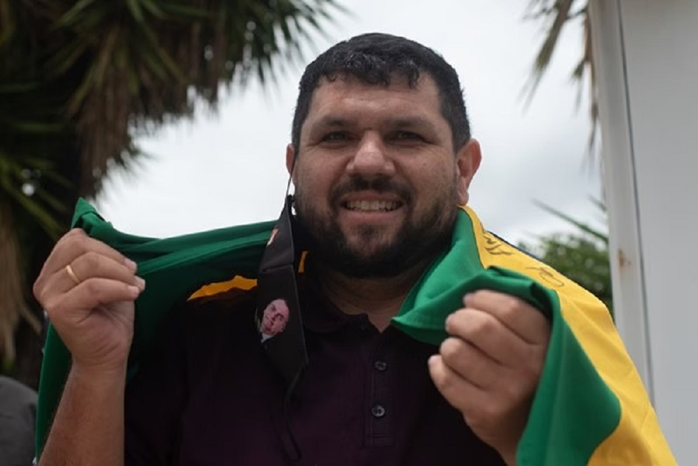 Oswaldo Eustáquio de camisa escura e com a bandeira do Brasil nas costas, falando