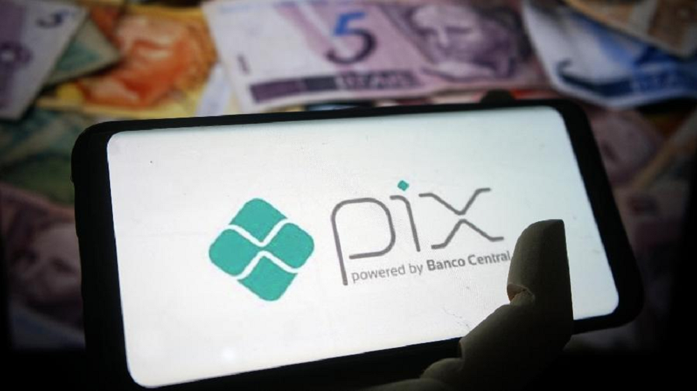 Mão segurando celular com tela com logo do Pix e cédulas de reais no fundo da foto