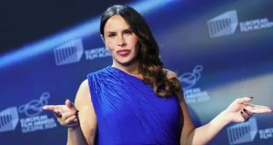 Karla Sofía Gascón de roupa azul, séria, com os braços abertos e cabelo solto