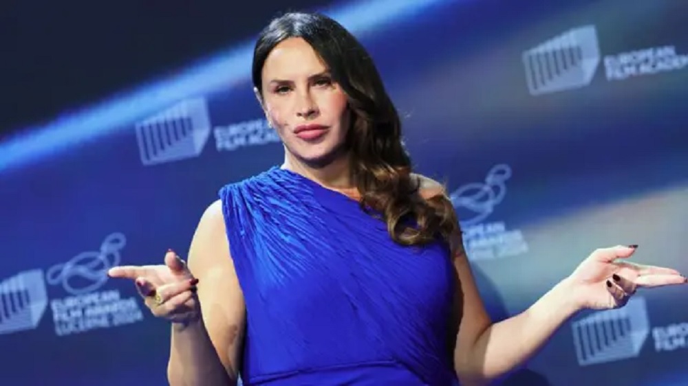 Karla Sofía Gascón de roupa azul, séria, com os braços abertos e cabelo solto