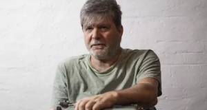 Marcelo Rubens Paiva de camiseta cinza clara, olhando pra frente, sério, sem olhar pra câmera