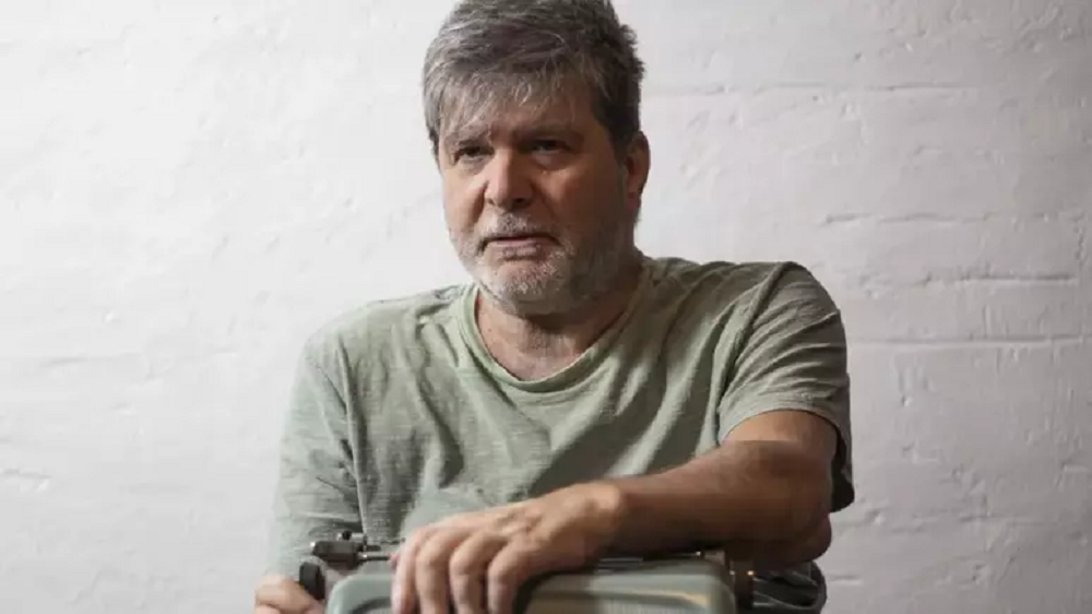 Marcelo Rubens Paiva de camiseta cinza clara, olhando pra frente, sério, sem olhar pra câmera