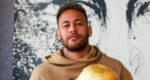 Neymar Jr. olhando para a câmera sem sorrir, de moletom bege, segurando bola de futebol dourada