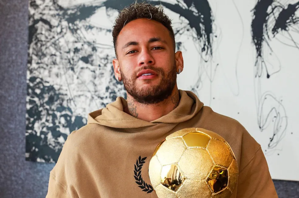 Neymar Jr. olhando para a câmera sem sorrir, de moletom bege, segurando bola de futebol dourada