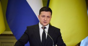 presidente da Ucrânia, Volodymyr Zelensky, falando em microfone, sério, com bandeiras ao fundo