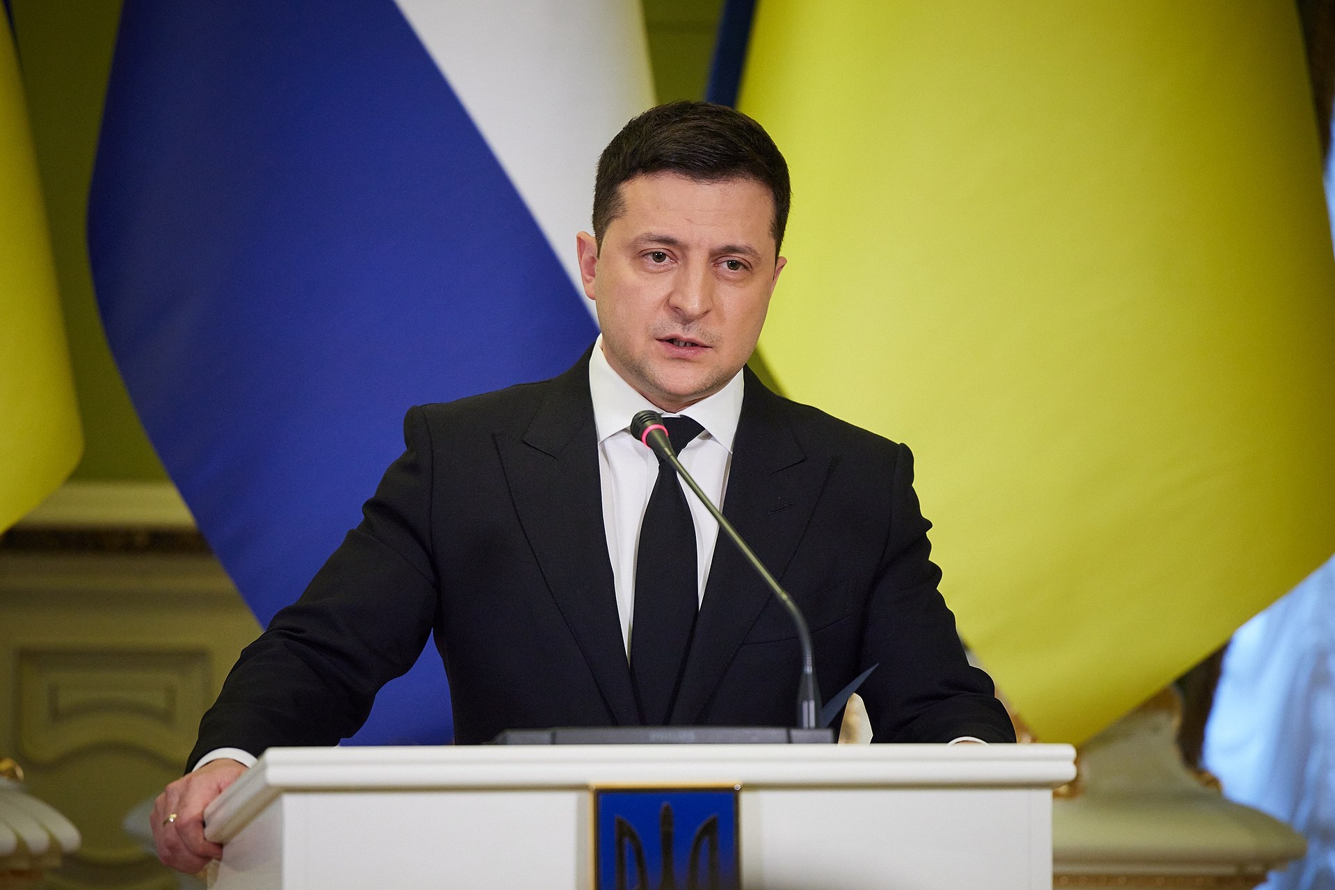 presidente da Ucrânia, Volodymyr Zelensky, falando em microfone, sério, com bandeiras ao fundo