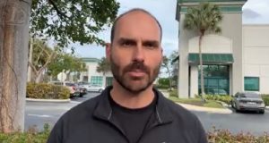 O deputado federal Eduardo Bolsonaro (PL-SP) de moletom cinza falando para a câmera com casas ao fundo