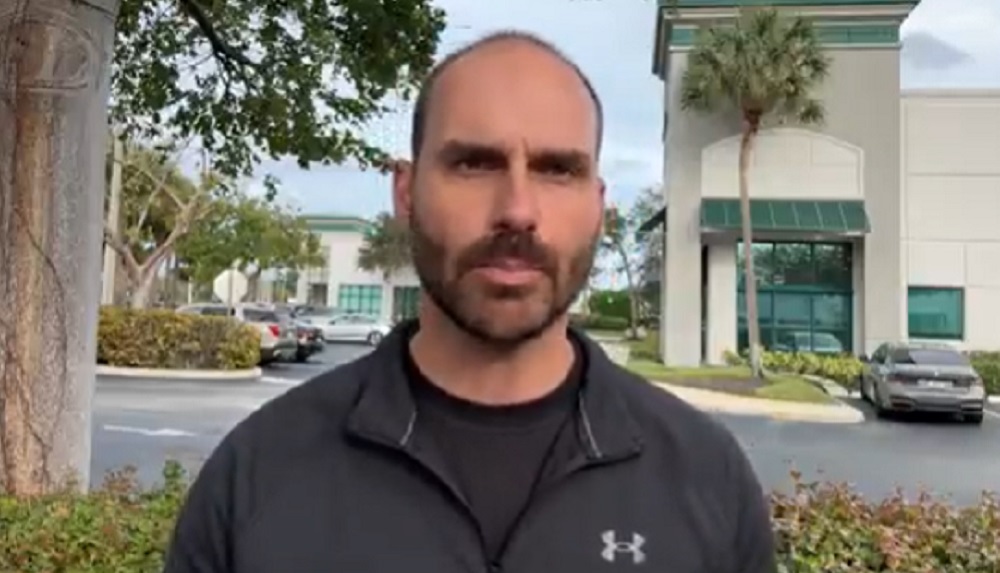 O deputado federal Eduardo Bolsonaro (PL-SP) de moletom cinza falando para a câmera com casas ao fundo