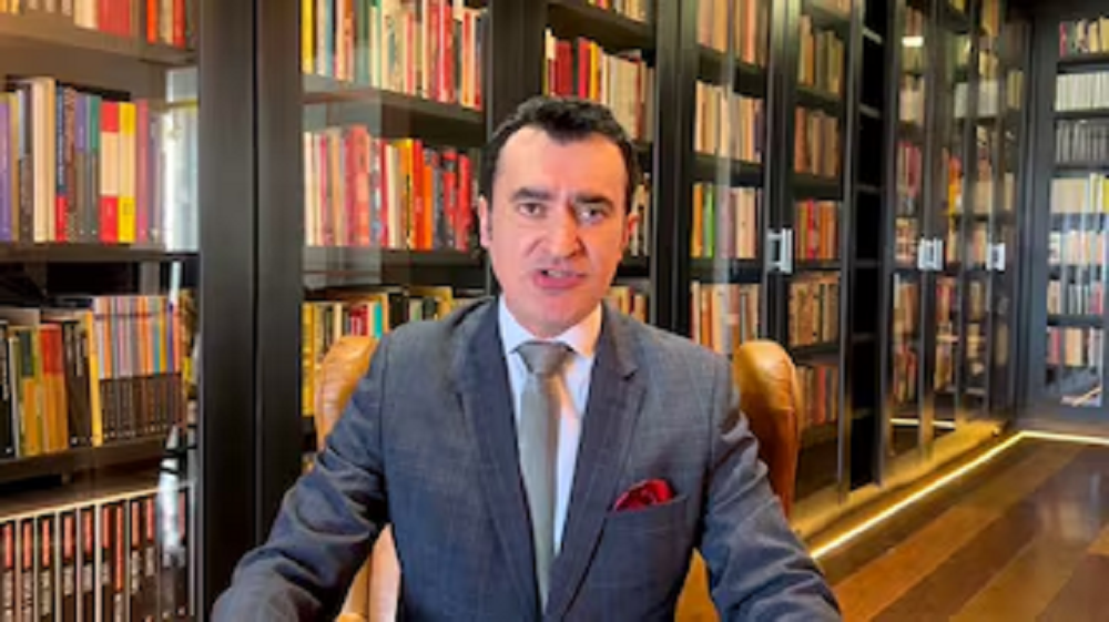professor Alysson Leandro Barbate Mascaro de terno e gravata, falando para a câmera perto de estante de livros