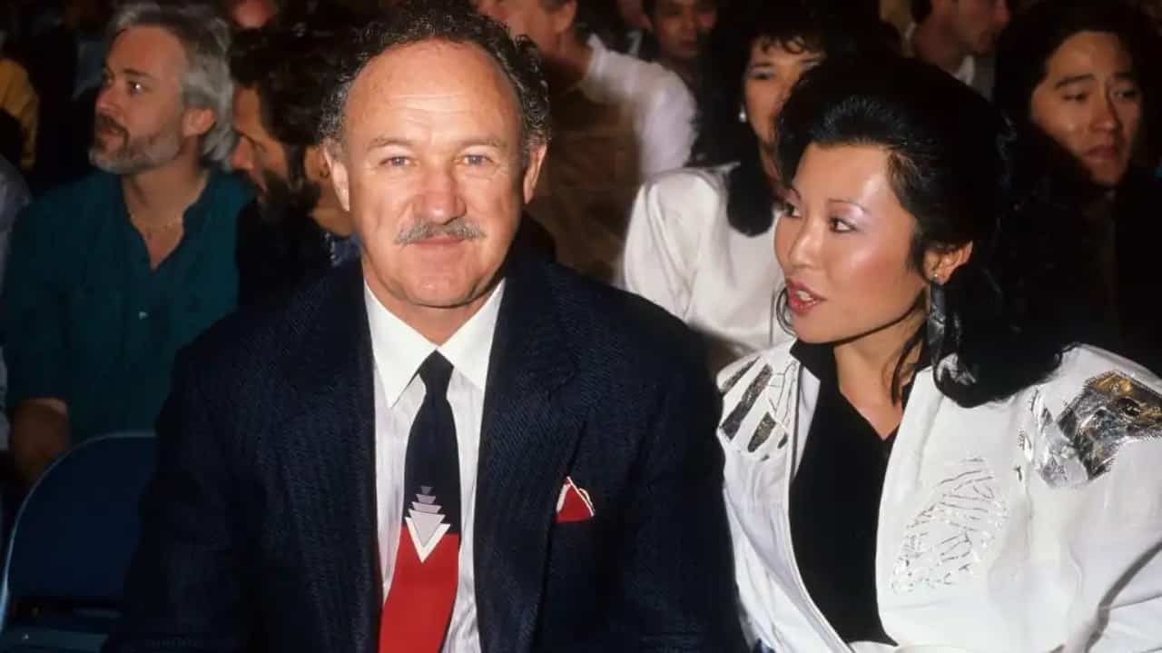Gene Hackman e Betsy Arawaka sentados lado a lado, ele sorrindo e ela olhando para ele e falando, em foto antiga