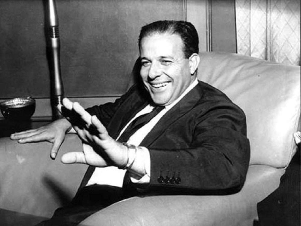 João Goulart sentado, em foto em preto e branco, sorrindo e acenando sem olhar para a câmera
