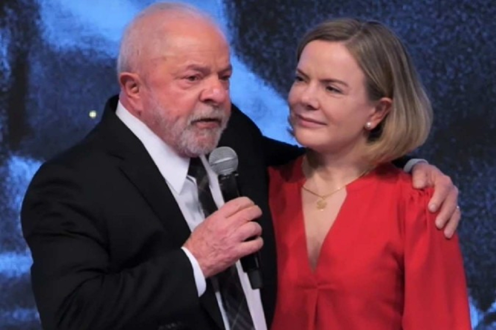Lula e Gleisi abraçados lado a lado, ele falando em microfone e ela olhando para ele