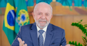 Lula sorrindo e gesticulando para a câmera com bandeiras do Brasil ao fundo