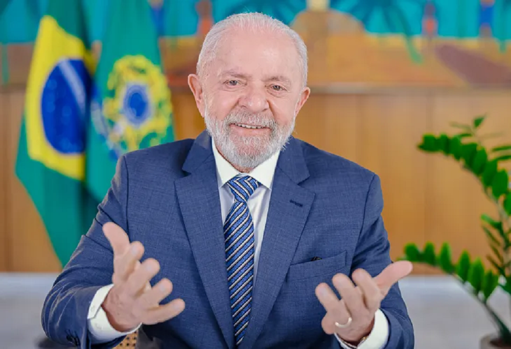 Lula sorrindo e gesticulando para a câmera com bandeiras do Brasil ao fundo
