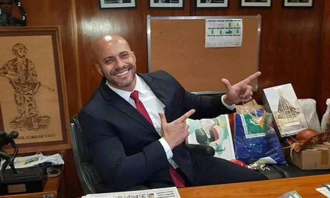 Daniel Silveira sorrindo e fazendo arminha com as mãos, sentado