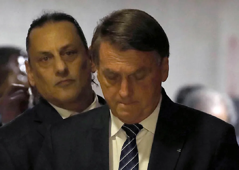 Frederick Wassef atrás de Bolsonaro, ambos sérios