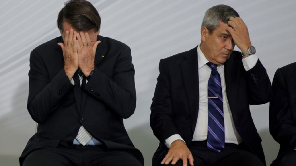 Jair Bolsonaro e Braga Netto lado a lado, ambos cabisbaixos com mãos no rosto