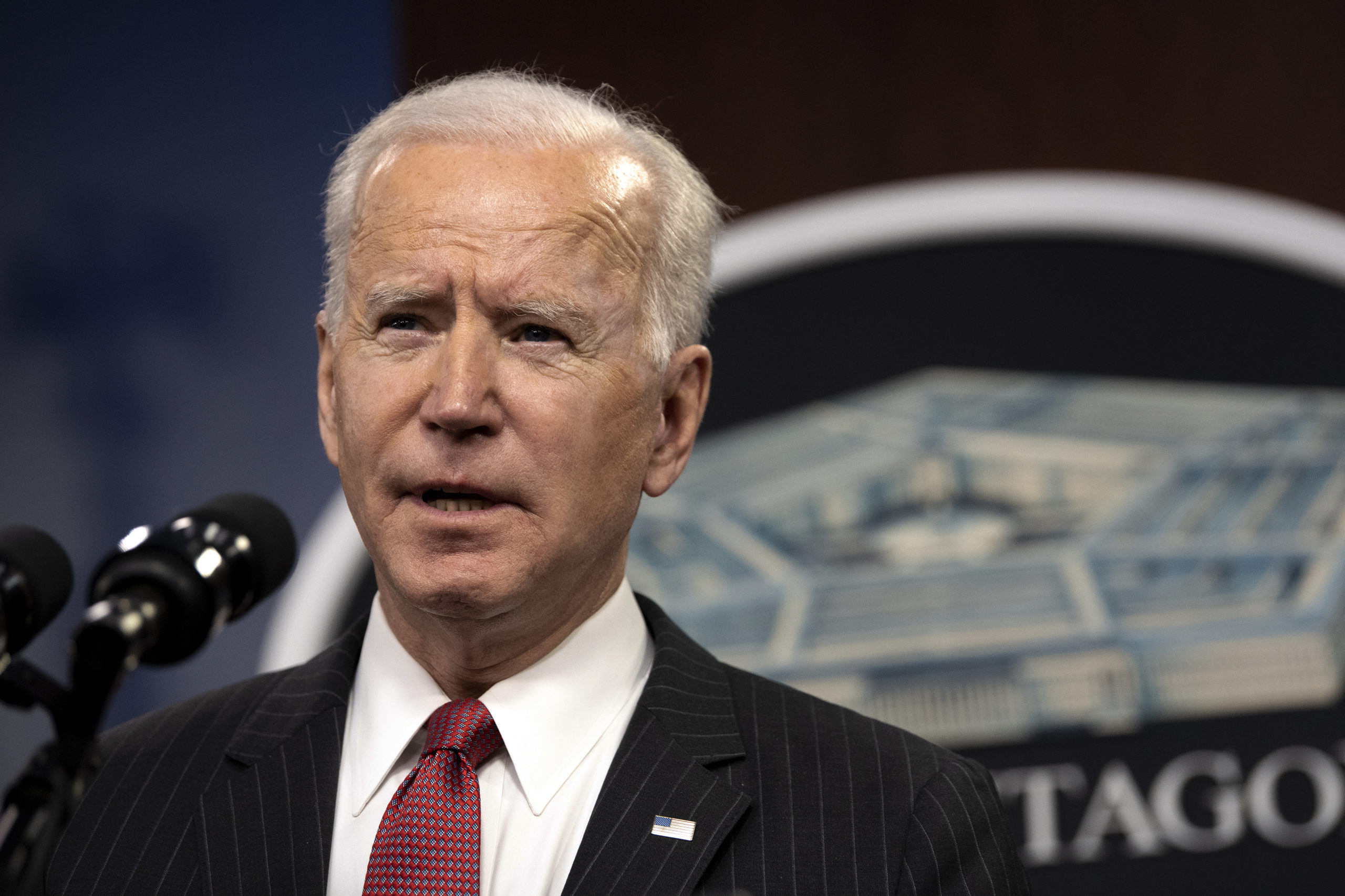 Joe Biden no canto esquerdo de foto, falando em microfone, sério