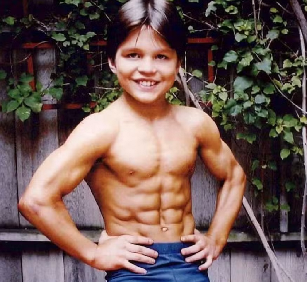 Richard Sandrak ainda criança, sem camisa, de sunga azul, sorrindo para a câmera