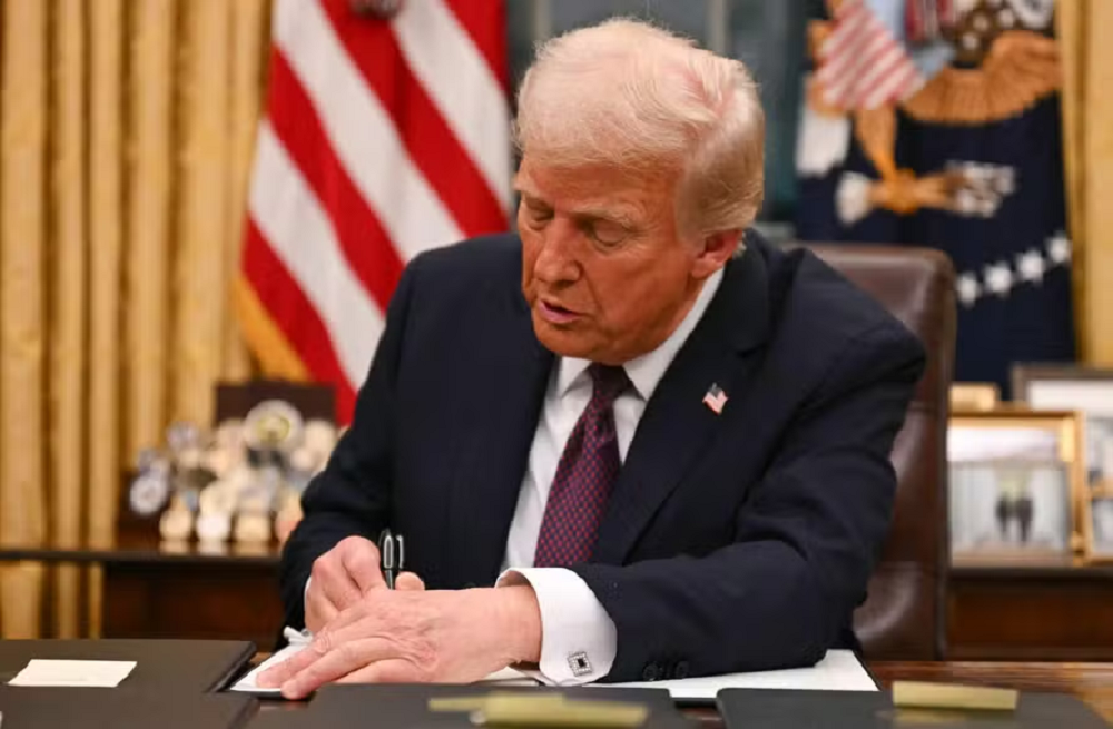 Donald Trump assinando documento na frente de bandeiras dos EUA