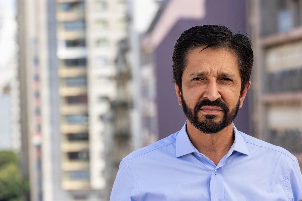 Ricardo Nunes de camisa social azul, com expressão séria e prédios ao fundo