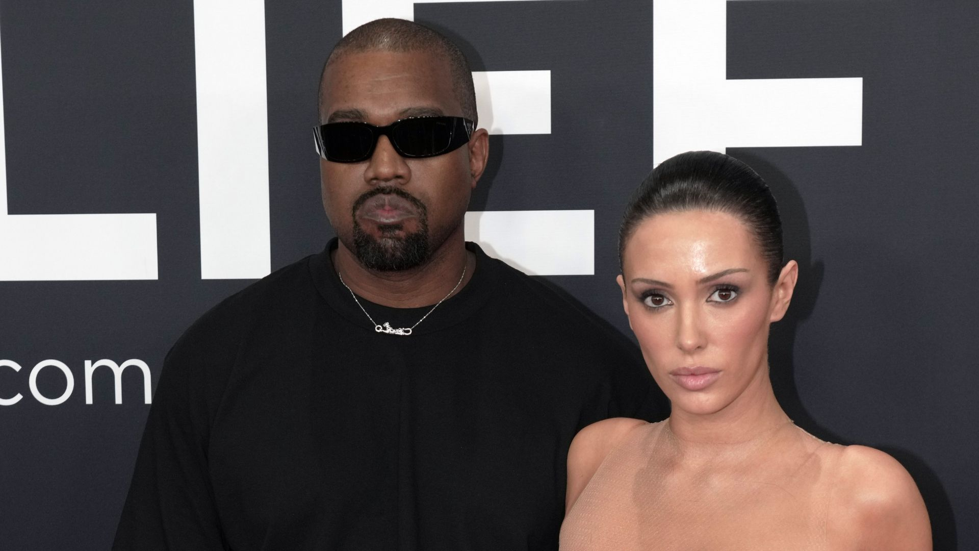 Kanye West de roupa preta e óculos escuros sério ao lado de Bianca Censori de cabelo solto e roupa transparente