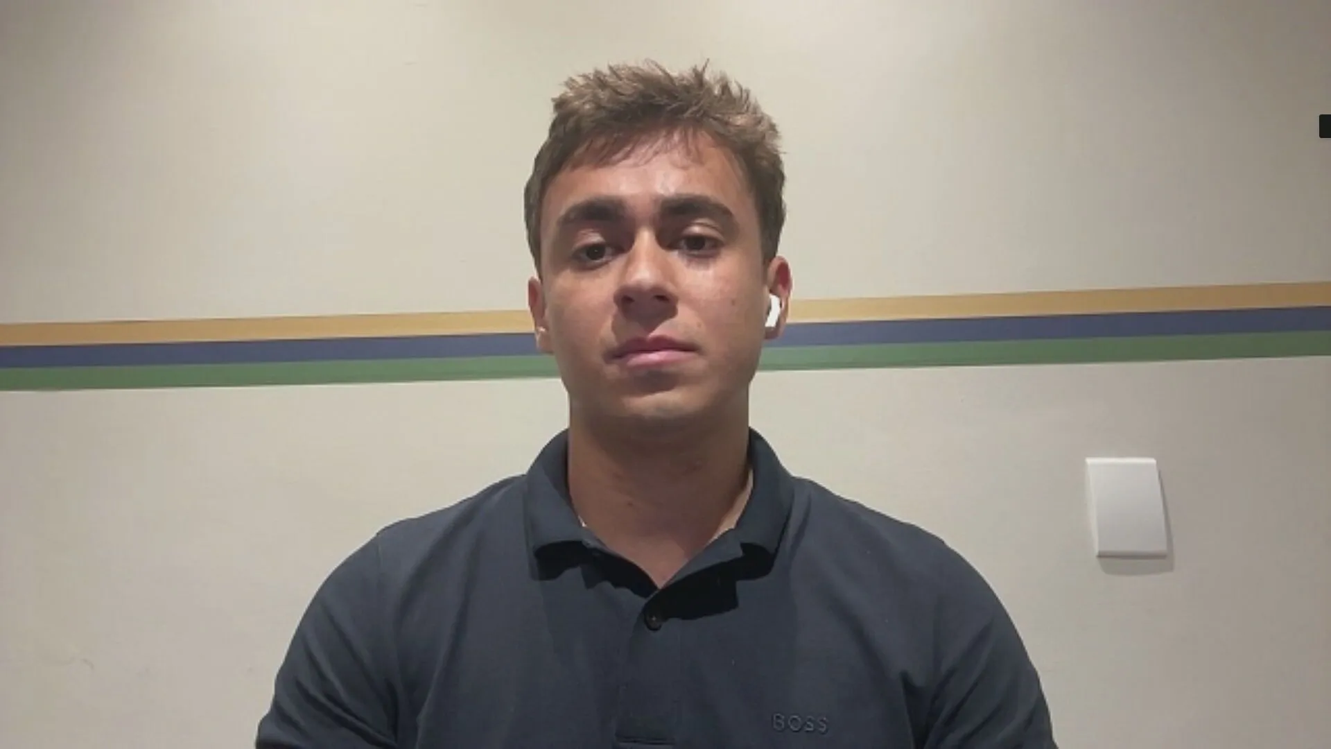 deputado federal Nikolas Ferreira (PL) de camisa polo preta olhando para a câmera, sério, com fone de ouvido branco sem fio