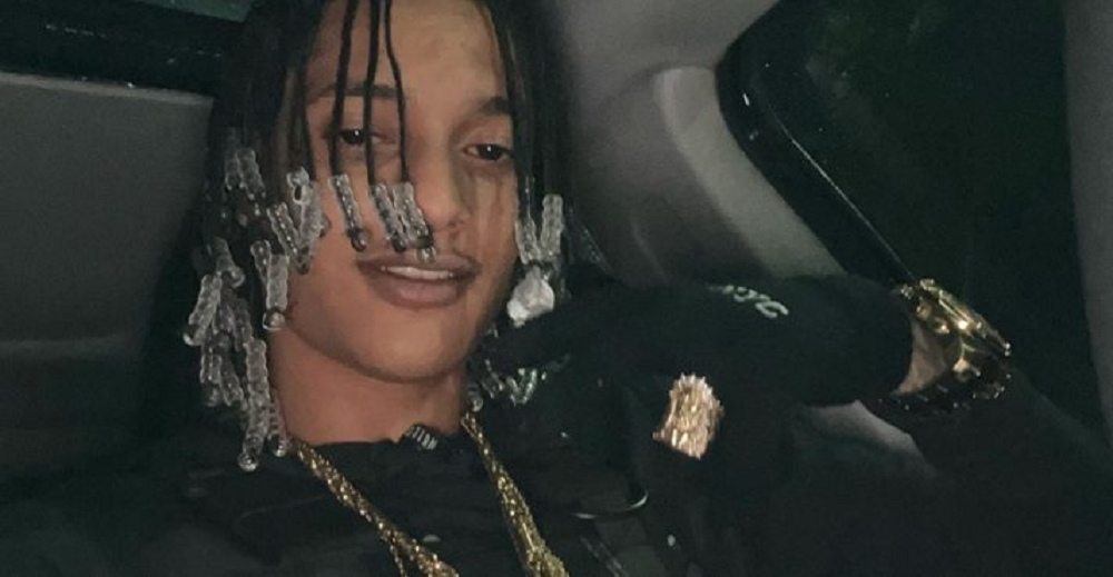 O rapper Oruam dentro de carro, de roupa e luvas pretas, sorrindo e gesticulando para a cãmera