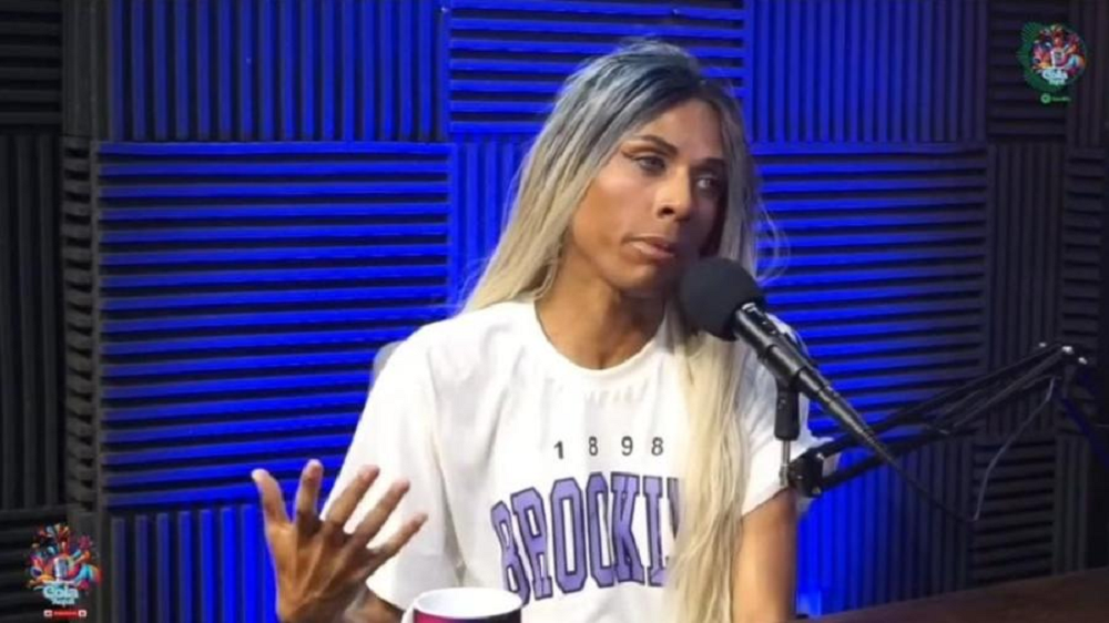 Brunella Hilson de camiseta branca, falando em microfone e gesticulando, de cabelo solto