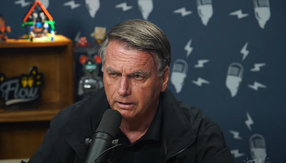 ex-presidente Jair Bolsonaro sério, falando em microfone, no Flow, de roupa de frio preta, sem olhar para a câmera
