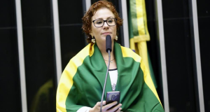 deputada federal Carla Zambelli (PL-SP) envolvida em bandeira do Brasil, segurando celular, séria, de óculos