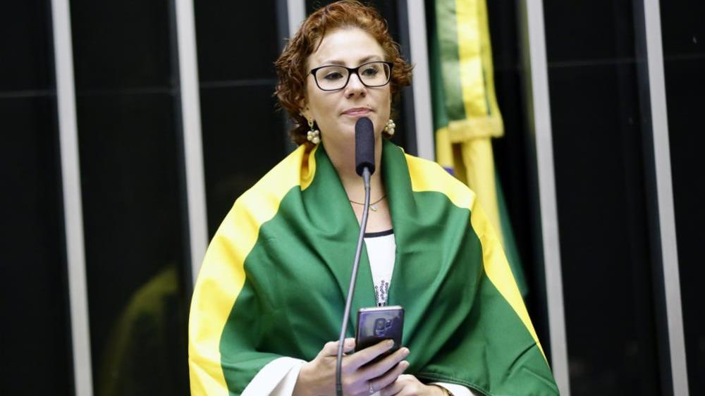 deputada federal Carla Zambelli (PL-SP) envolvida em bandeira do Brasil, segurando celular, séria, de óculos