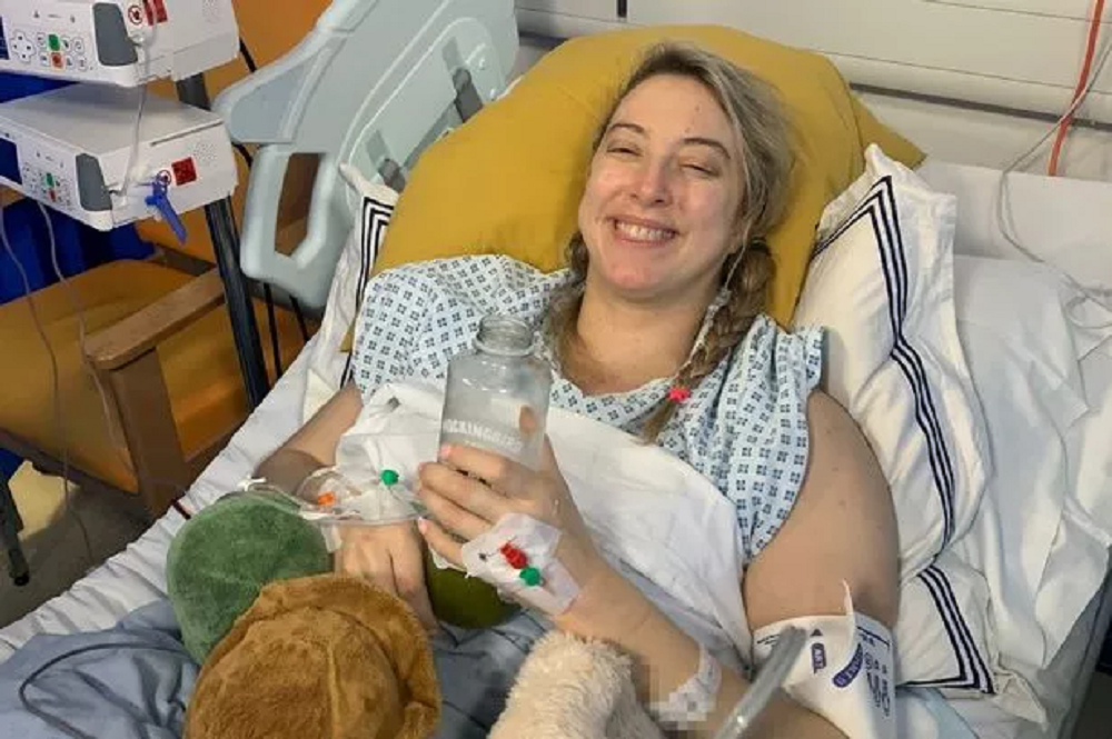 Abi Feltham deitada em cama de hospital, sorrindo e segurando garrafa