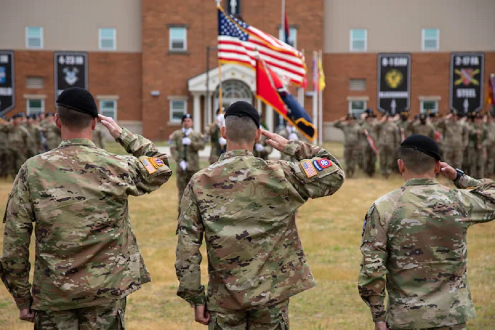 Militares do Exército de costas, batendo continência, perto de bandeira dos EUA
