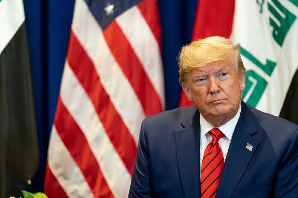 Donald Trump no canto direito de foto, sério, sem olhar para a câmera, com bandeira dos EUA ao fundo