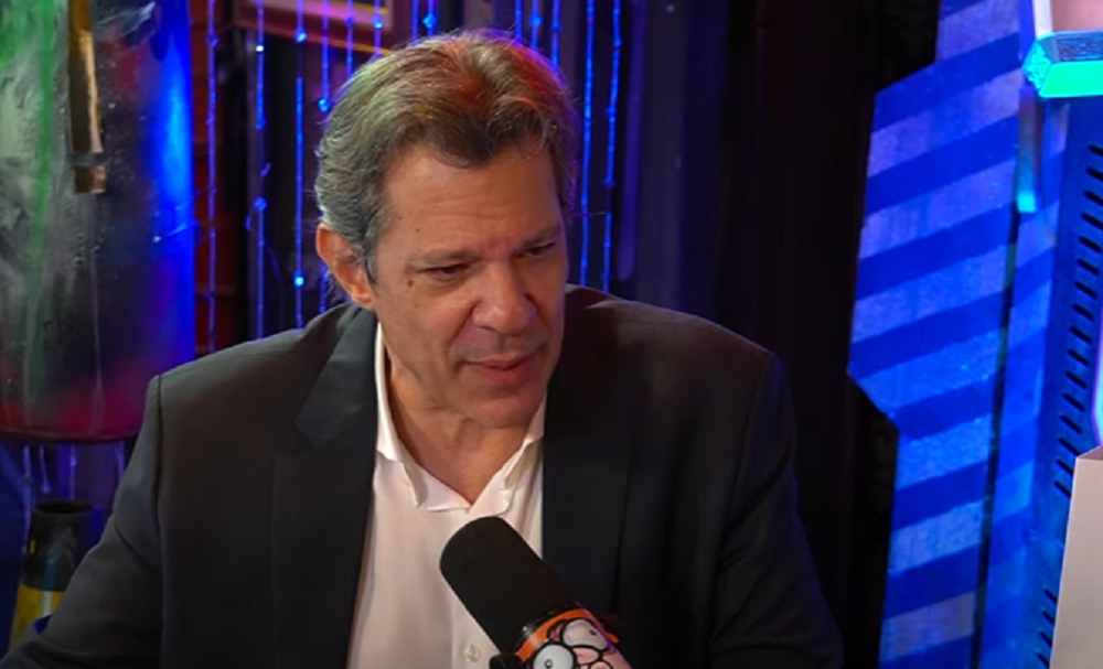 O ministro da Fazenda, Fernando Haddad, de camisa branca e paletó preto, falando, sério, sem olhar para a câmera