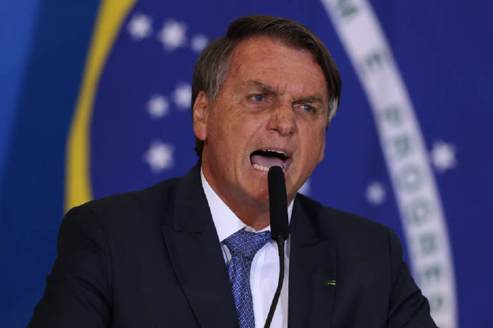 ex-presidente Jair Bolsonaro (PL) gritando em microfone, com bandeira do Brasil ao fundo