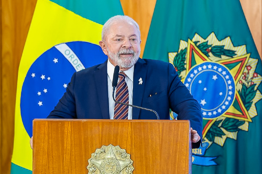 O presidente Luiz Inácio Lula da Silva (PT) sorrindo, com bandeiras ao fundo, em pé, sem olhar para a câmera