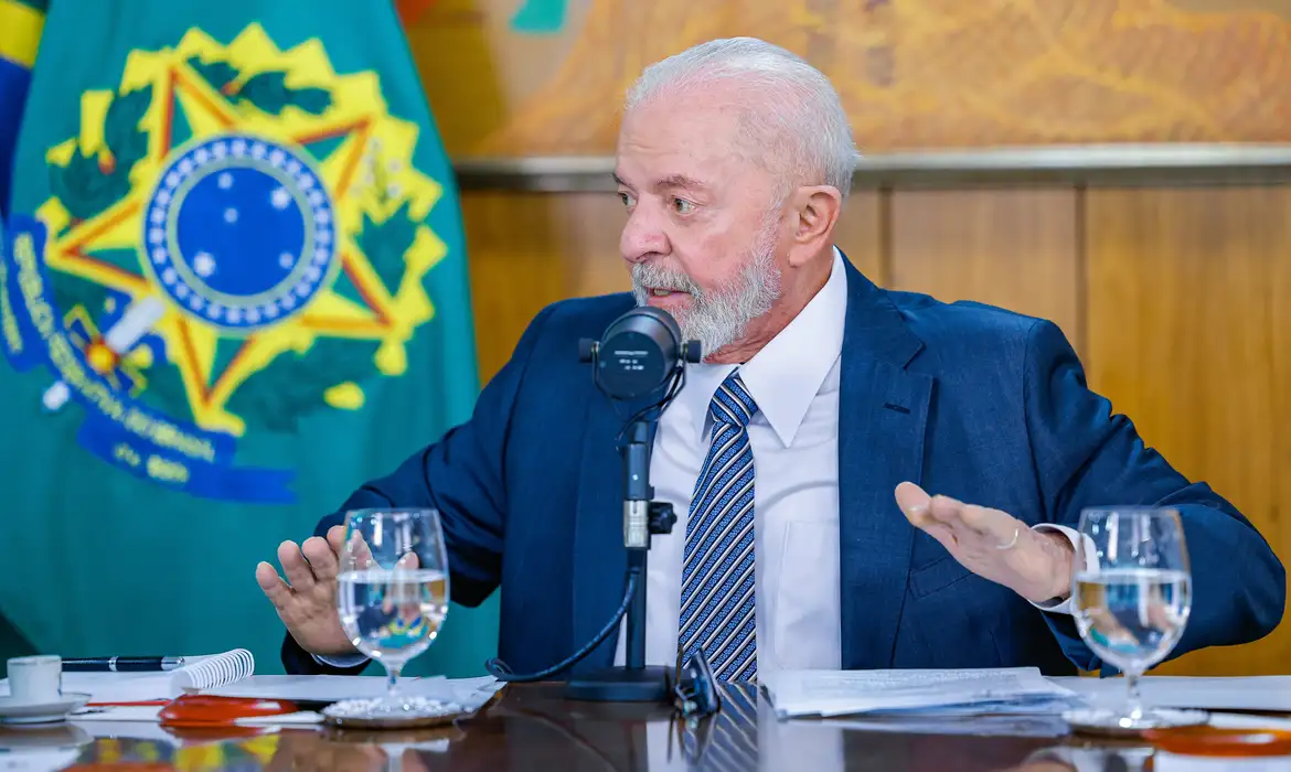 O presidente Luiz Inácio Lula da Silva falando, sentado à mesa, olhando para o lado