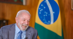Lula sorrindo no canto esquerdo da foto, com bandeira do Brasil ao fundo