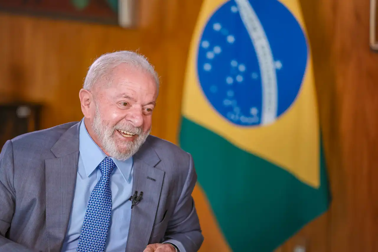 Lula sorrindo no canto esquerdo da foto, com bandeira do Brasil ao fundo