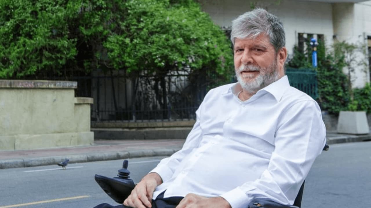 Marcelo Rubens Paiva de roupa social branca, com sorriso tímido para foto, com árvores e casa ao fundo