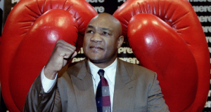 ex-pugilista George Foreman entre luvas de boxe gigantes, olhando pra cima com punho fechado, de terno bege e gravata colorida