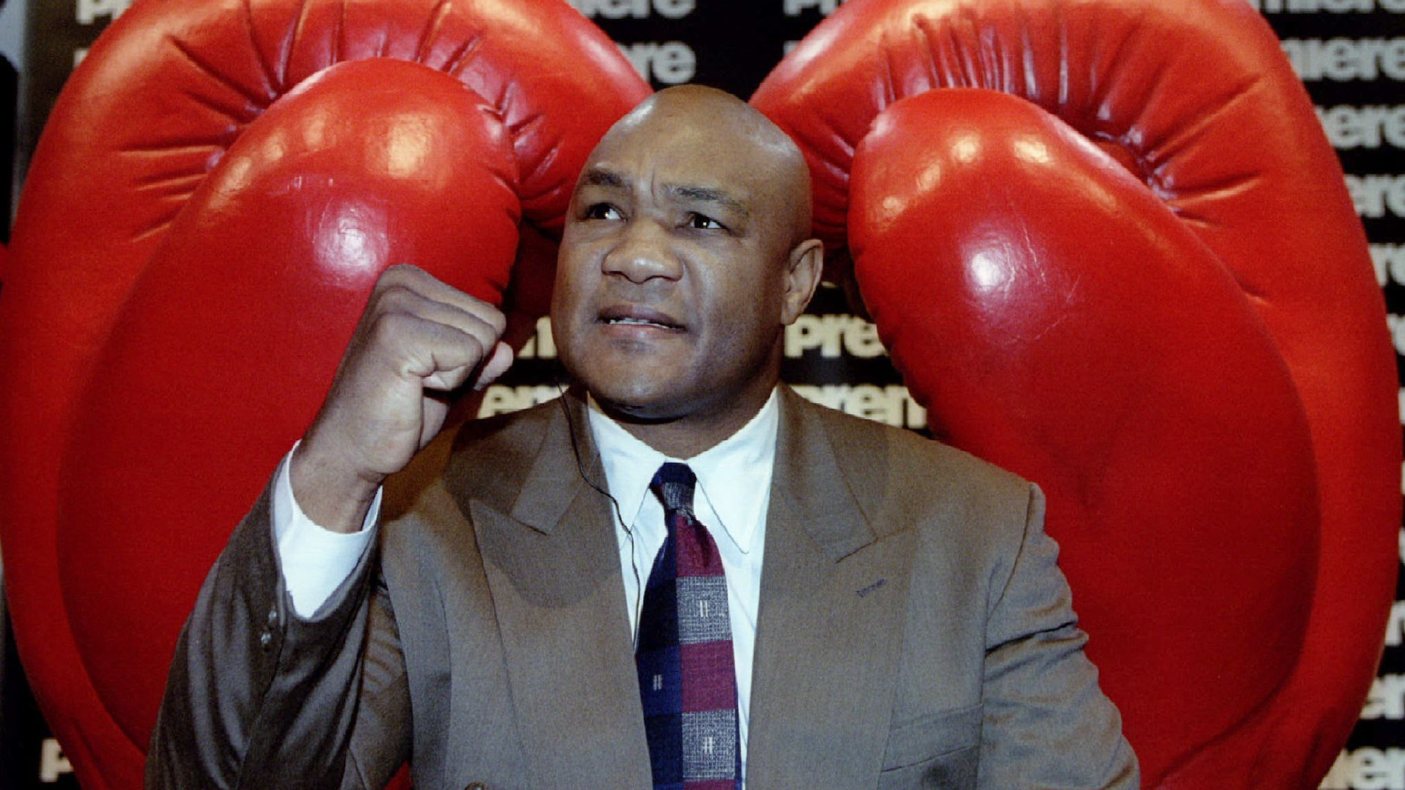 ex-pugilista George Foreman entre luvas de boxe gigantes, olhando pra cima com punho fechado, de terno bege e gravata colorida