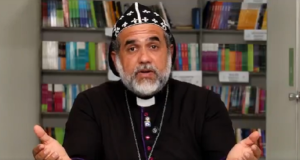 O padre Kelmon Luis Souza falando para a câmera, sério, com livros ao fundo