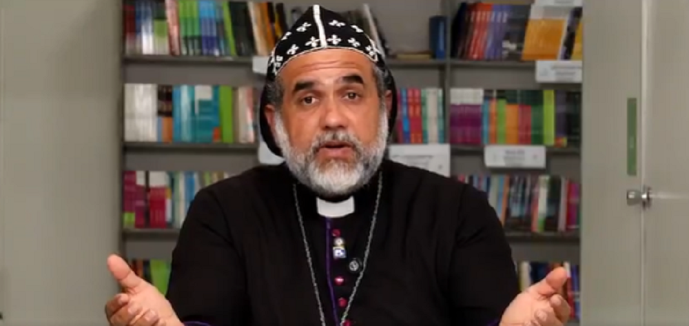 O padre Kelmon Luis Souza falando para a câmera, sério, com livros ao fundo