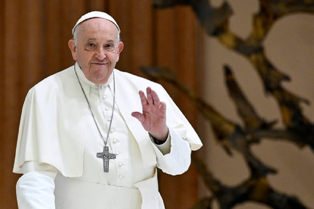 Papa Francisco em pé, acenando, com sorriso tímido, olhando para a câmera