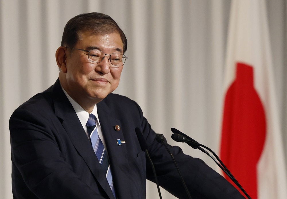 O primeiro-ministro japonês, Shigeru Ishiba, sorrindo de maneira tímida, perto de microfone e de bandeira do Japão